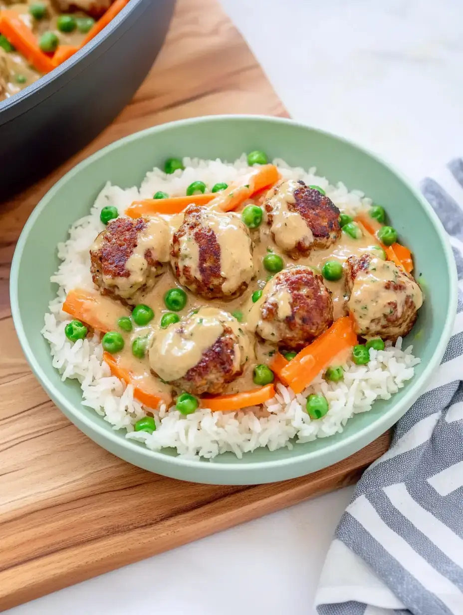 Une assiette avec du riz, des boulettes de viande dans une sauce crémeuse, des petits pois et des lamelles de carottes, servie dans un bol vert.