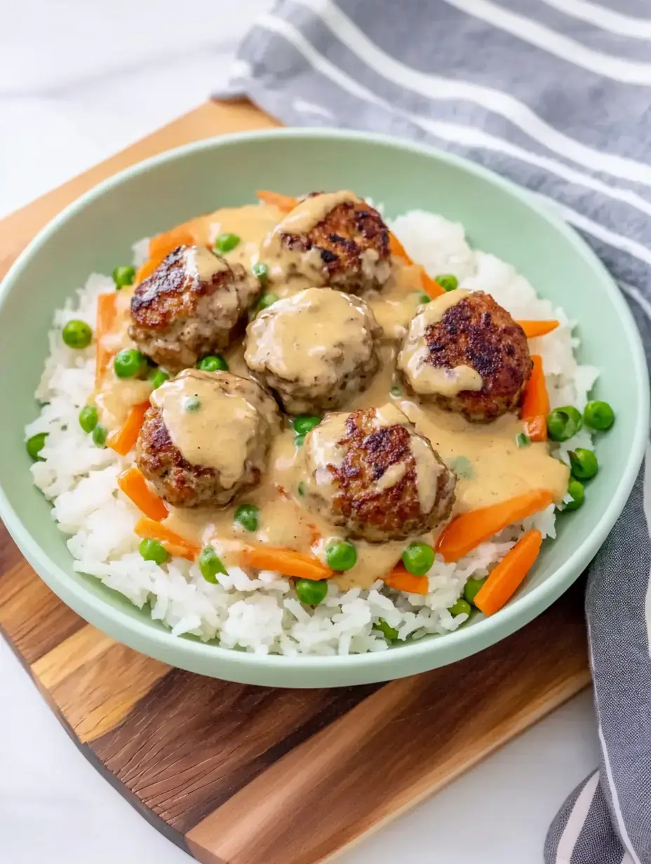 Un bol de riz garni de boulettes de viande dans une sauce crémeuse, entouré de petits pois et de lamelles de carottes.