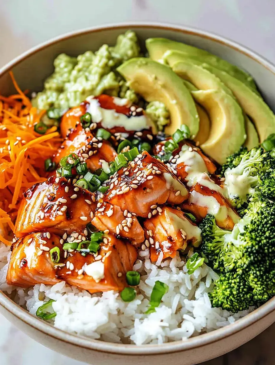 Un bol rempli de riz, saumon grillé, tranches d'avocat, brocoli, lamelles de carottes et sauce crémeuse, garni de sésame et oignons verts.