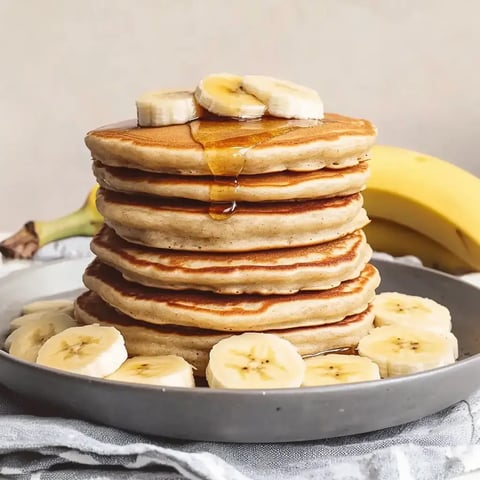 Une assiette avec une haute pile de crêpes, garnie de tranches de banane et de sirop, posée à côté d'une banane.
