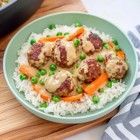 Une assiette de riz avec des boulettes de viande dans une sauce crémeuse, garnie de carottes et petits pois.