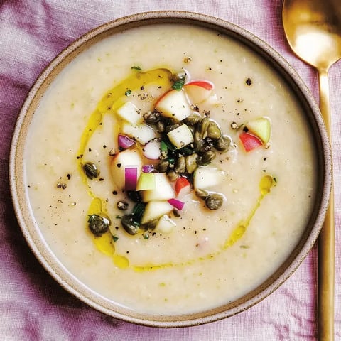 Un bol de soupe crémeuse, garnie de petits morceaux de pomme, câpres et un filet d'huile d'olive, sur un tissu rose.