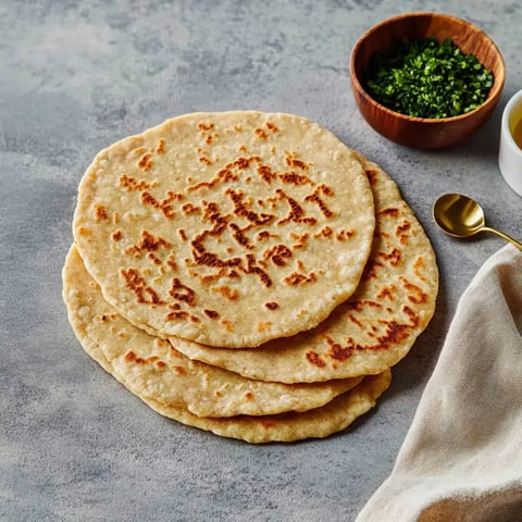 Une pile de galettes fraîchement cuites repose sur une surface grise marbrée, accompagnée d'un bol de verdure hachée.