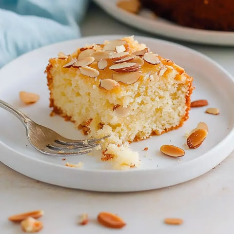 Une part de gâteau aux amandes sur une assiette blanche, garnie d'amandes effilées, posée à côté d'une fourchette.