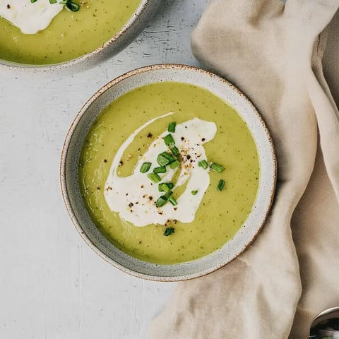 Deux bols de soupe verte, garnis de crème, ciboulette et poivre, posés sur une surface claire.