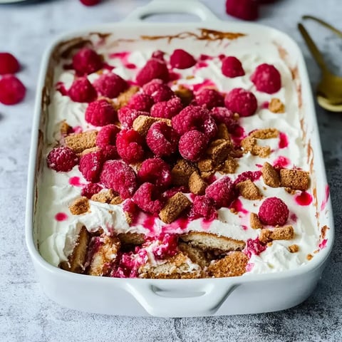 Un dessert dans un plat blanc, garni de crème, framboises et miettes de biscuits.