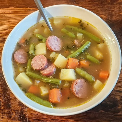 Un bol de soupe aux légumes contenant des saucisses, haricots verts, carottes et morceaux de pommes de terre.