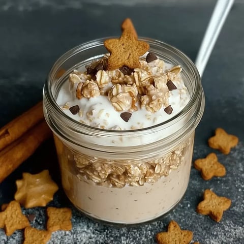 Un pot contenant des flocons d'avoine, du yaourt et des morceaux de chocolat, décoré d’un biscuit étoilé, entouré de bâtons de cannelle et de petits biscuits.