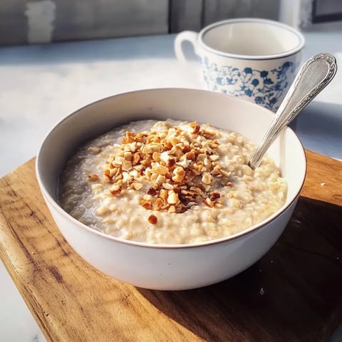 Un bol de porridge garni de noix hachées, posé sur une planche en bois à côté d'une tasse.